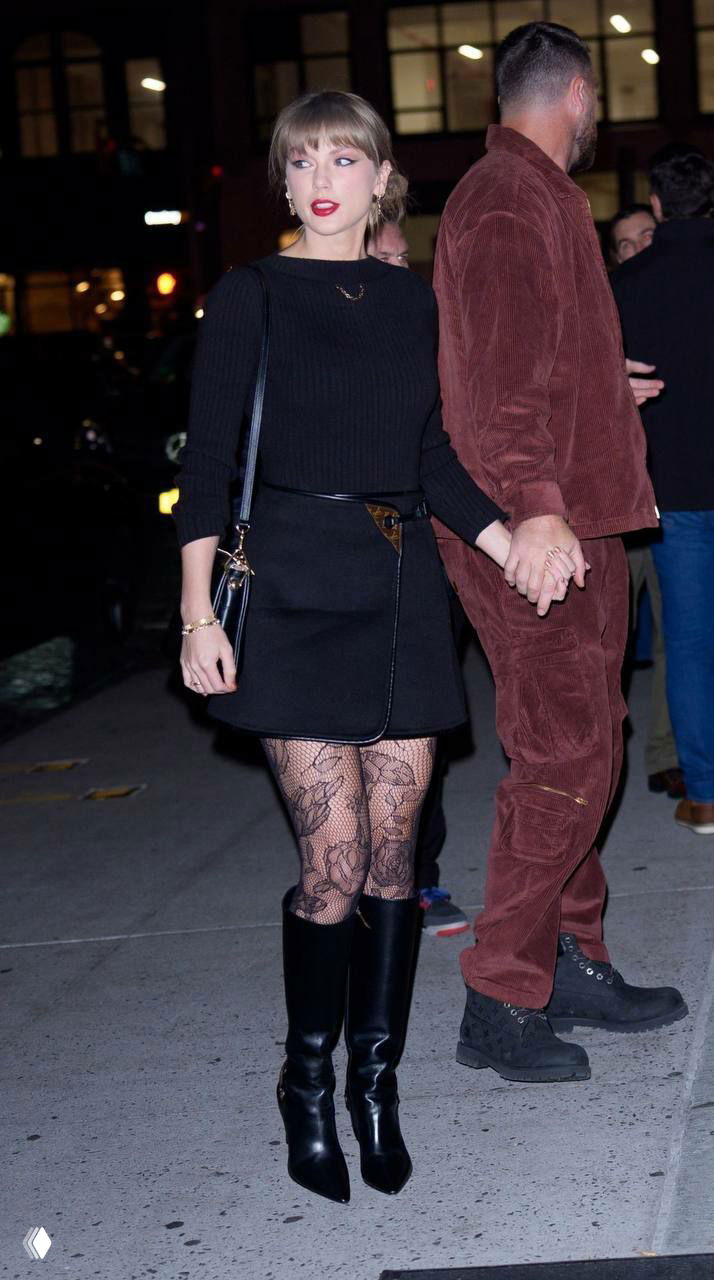 Ночной папарацци-кадр: женщина в чёрном платье и высоких сапогах идёт по тротуару рядом с мужчиной, городские огни ночью.