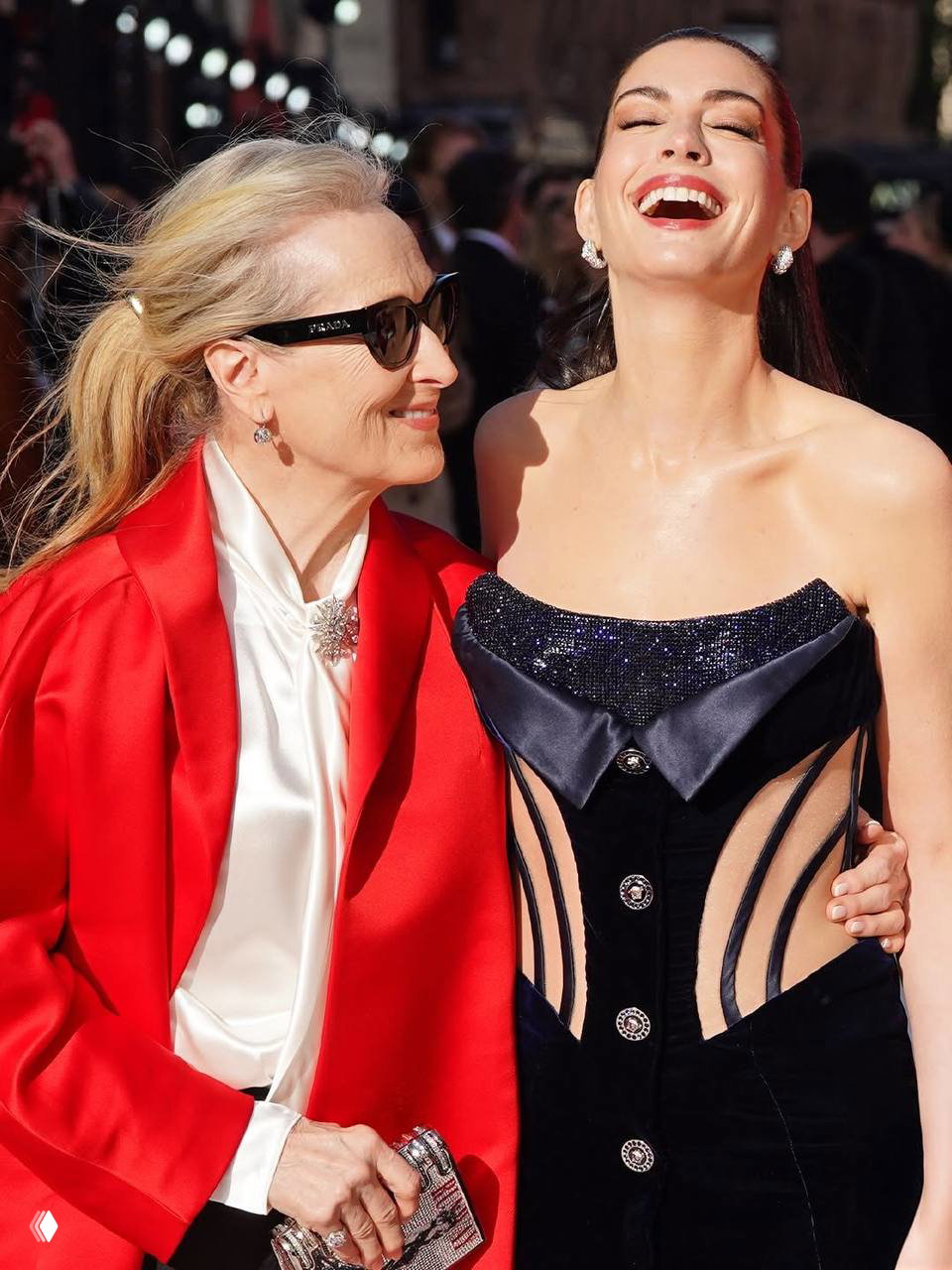 Две женщины смеются на дневной красной дорожке: одна в ярком красном пальто, другая — в тёмном корсетном платье; солнечный свет подчёркивает образы.