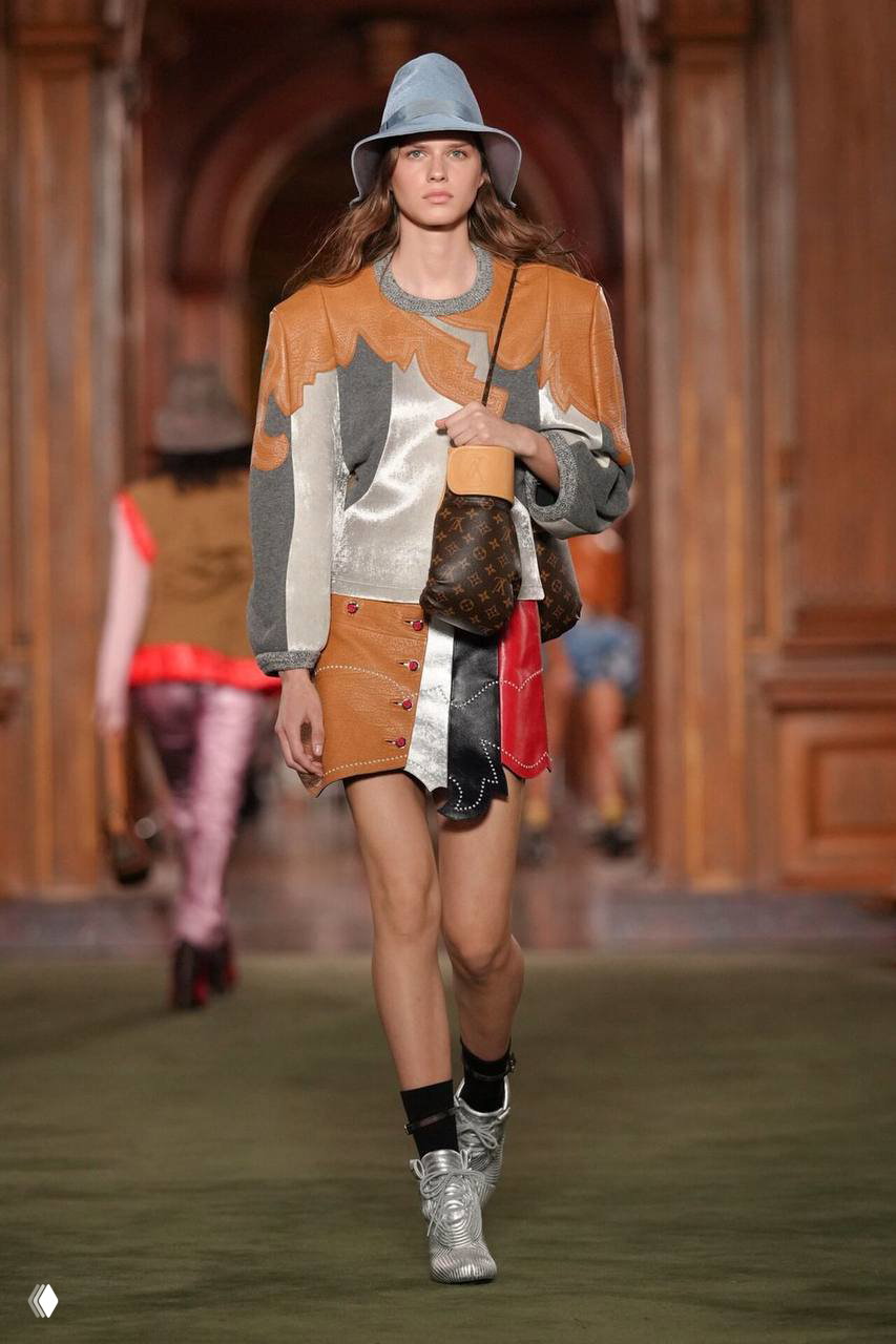 Асимметричный наряд в технике патчворк с мини-юбкой и сумкой Louis Vuitton с монограммой идёт по подиуму в музее Фрика во время шоу 2027 года.