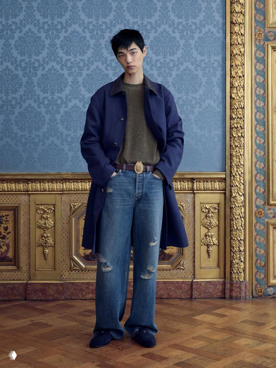 Модель в свободных джинсах и коричневом пиджаке, с сумкой через плечо, позирует у золочёной панели и картины — деним и дворянская эстетика в кадре.