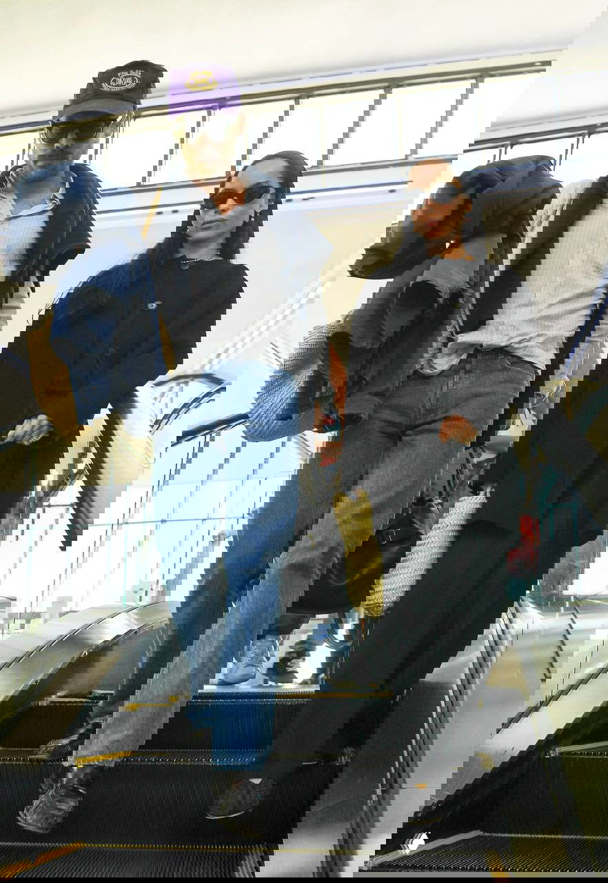 Фото пары в кадре эскалатора: Зои в чёрном пальто и сумке‑корзине, Гарри в джинсах и кепке — кадр подчёркивает их согласованную эстетику путешествий и улицы.