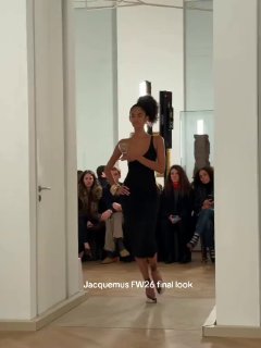 Модель на подиуме в черном асимметричном платье, держит бокал у груди; финальный выход показа, зрители на заднем плане.