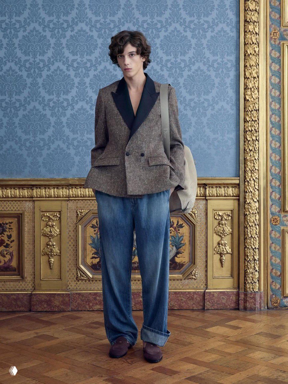 Модель в тёмном пальто и широких потертых джинсах, с акцентом на ремень с большой пряжкой, на фоне декоративного панно и паркетного пола XVII‑векового интерьера.