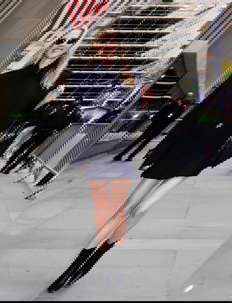 Городской кадр: женщина в тёмном пальто шагает по тротуару, на заднем плане ограждение, строительные леса и национальный флаг.