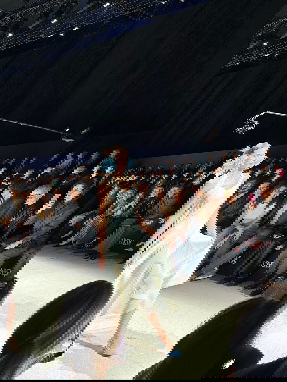 Модель в лёгком белом платье с голубыми аксессуарами на показе Leffers; передние ряды публики наблюдают и некоторые снимают на телефоны.