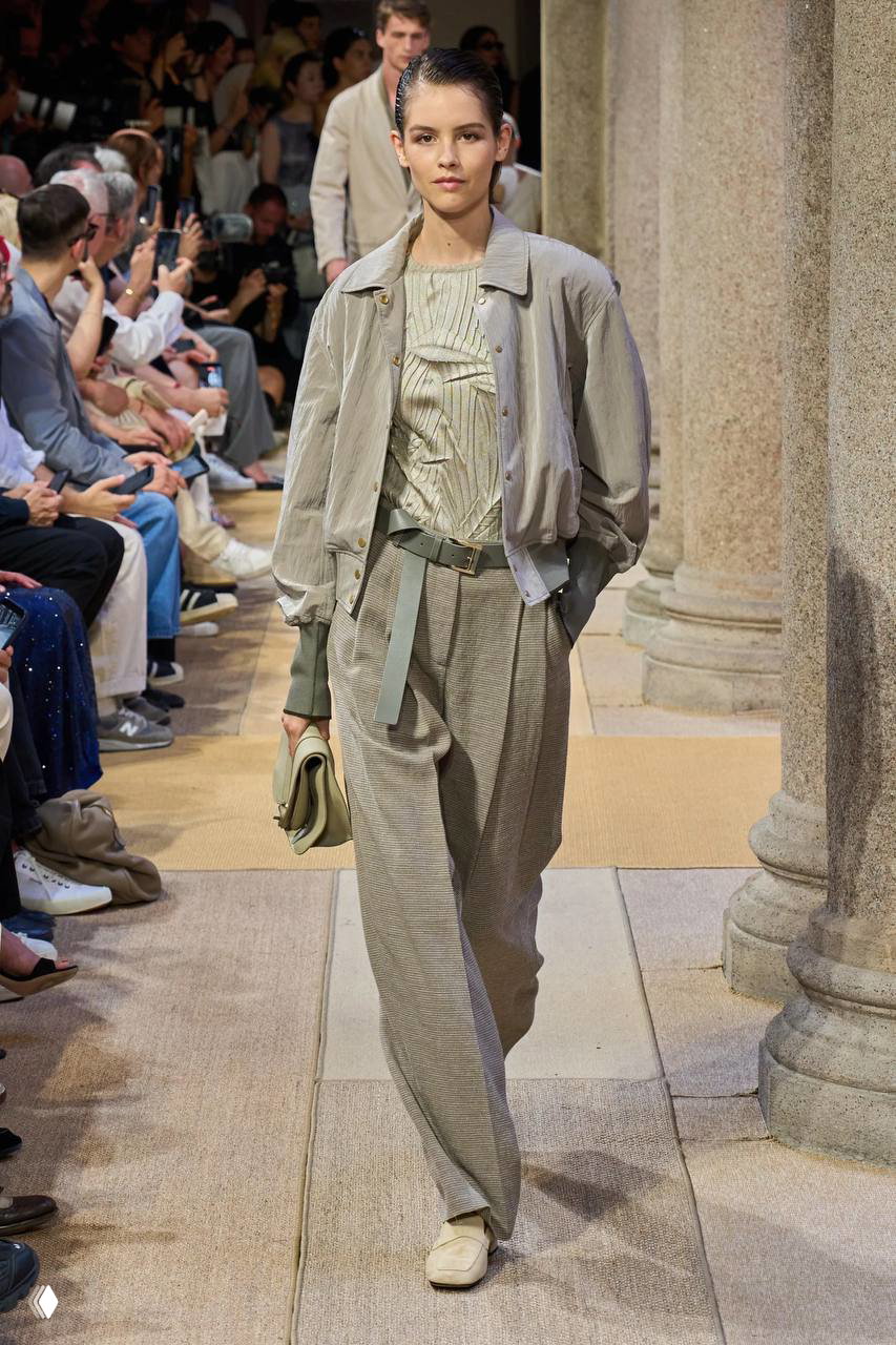 Женская модель в светлой куртке и широких брюках идёт по подиуму Giorgio Armani; пояс, клатч и лёгкая фактура тканей заметны на первом плане.