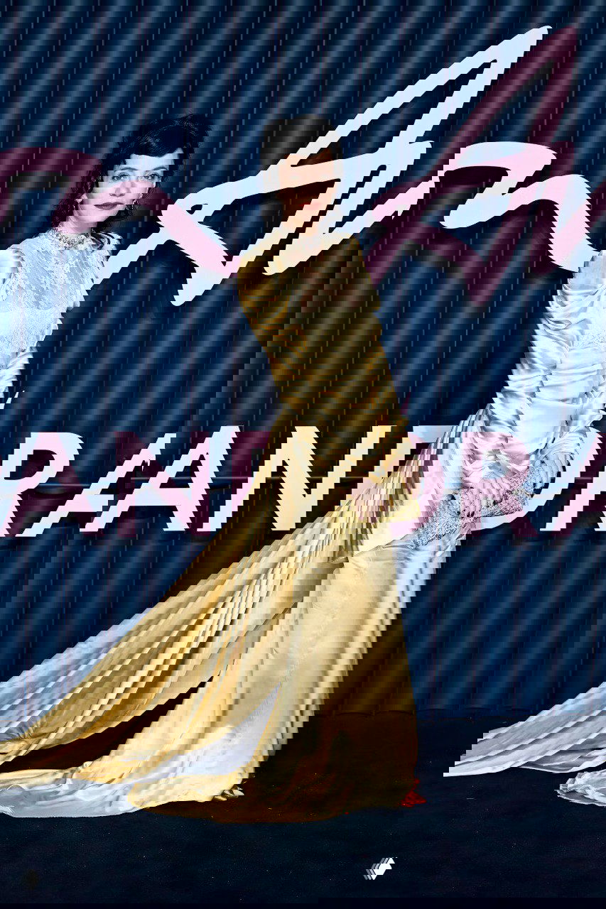 Женщина в золотистом атласном платье с кружевной вставкой и длинным шлейфом позирует у брендированной синей стены с розовой надписью.