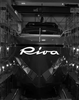 Чёрно‑белый кадр носовой части яхты Riva с крупным логотипом на корпусе, судно стоит в ангаре или доке на фоне оборудования.