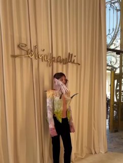 Фотозона с брендовым логотипом Schiaparelli и человеком в ярком наряде с бахромой, кадр демонстрирует подготовку и стилистические детали шоу.
