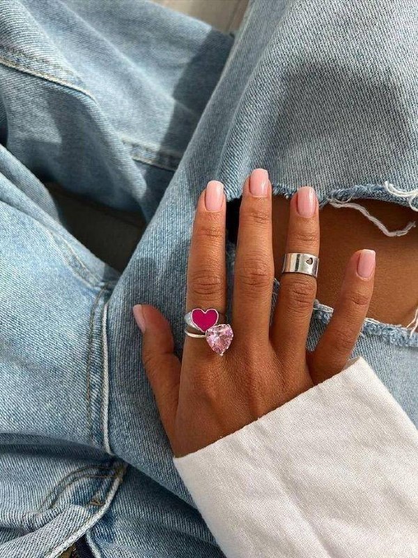 Рука с короткими ногтями в светло-розовом/нюдовом тоне на фоне джинсовой ткани; аккуратный полупрозрачный маникюр и декоративные кольца.