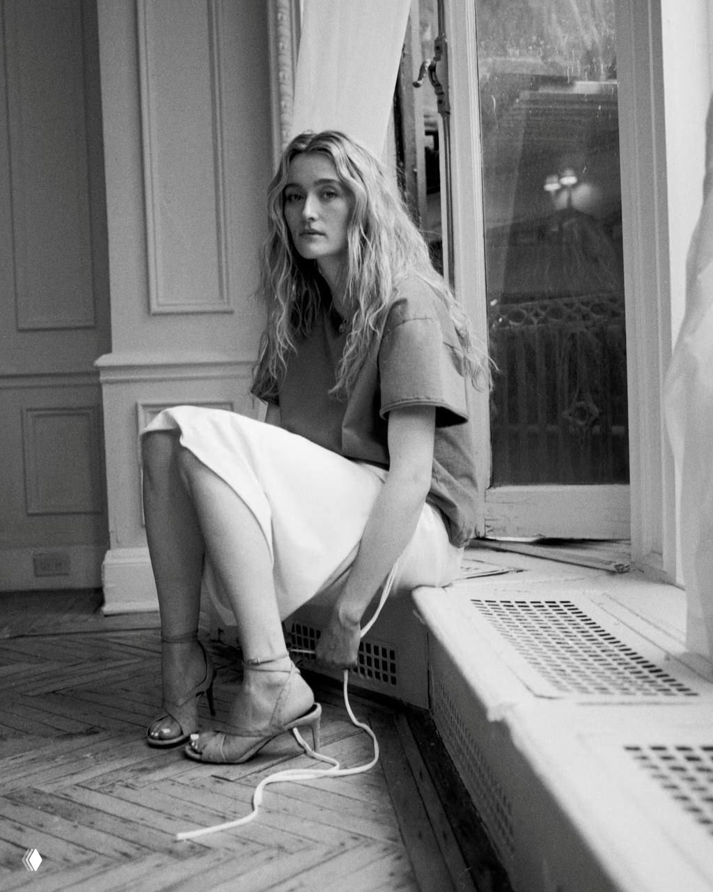Чёрно‑белый портрет Сары Пиджон у окна в повседневном образе: футболка и юбка, спокойная интерьерная съёмка для NYLON.