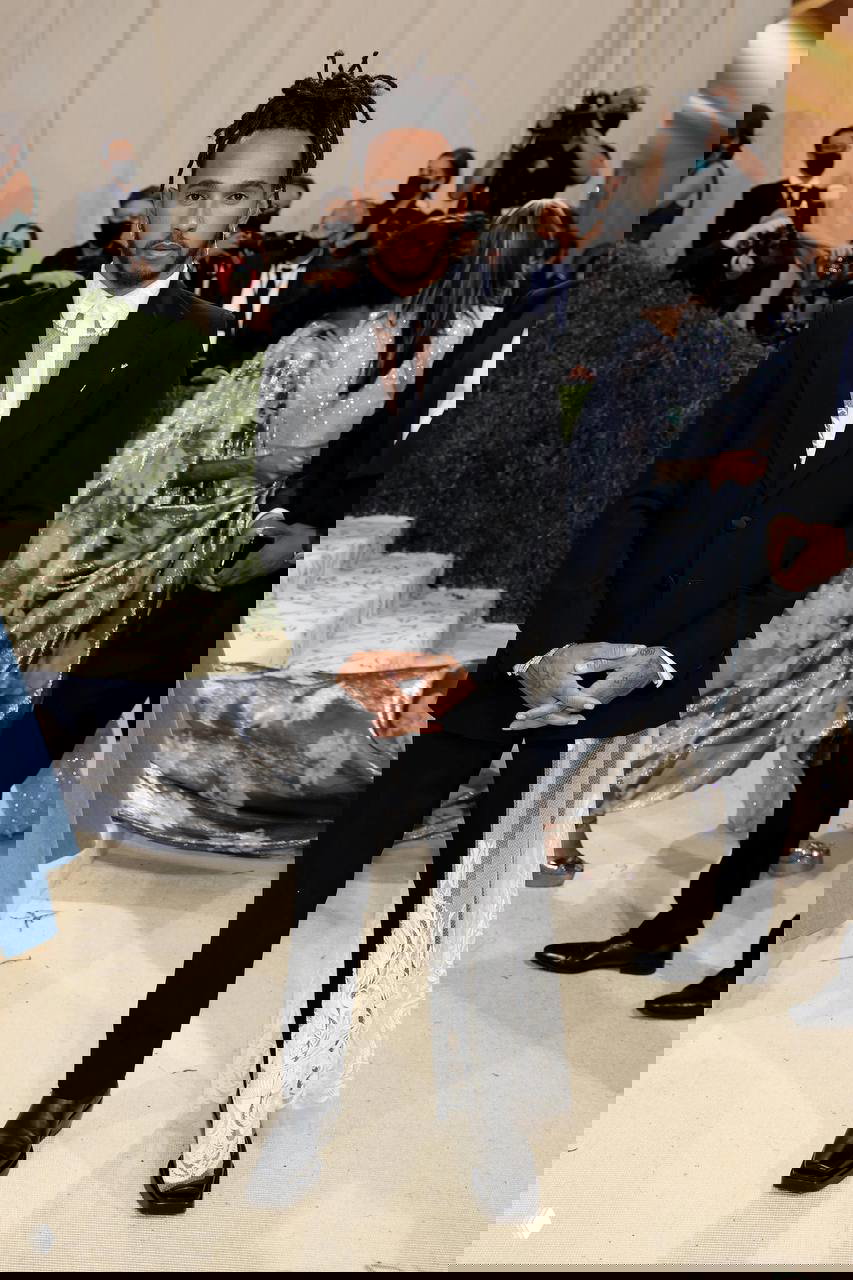 Льюис Хэмилтон в черном костюме с прозрачной деталью и воздушным шлейфом на фоне камер; вечерний образ Met Gala и внимание прессы.