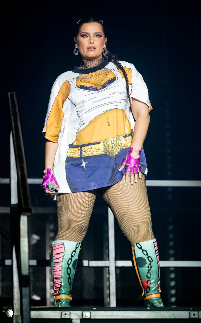 Крупный план Нелли Фуртадо в сценическом костюме и сеточных колготках: серьёзное лицо, яркие аксессуары и концертный свет вокруг неё.