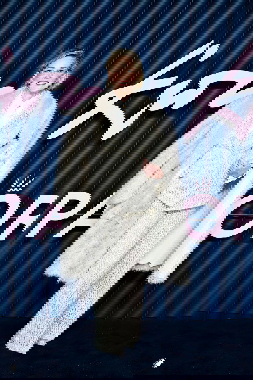 Женский образ в светлом фактурном костюме и длинной меховой шубе, улыбающаяся позирует у фирменного синего фона с розовым логотипом.