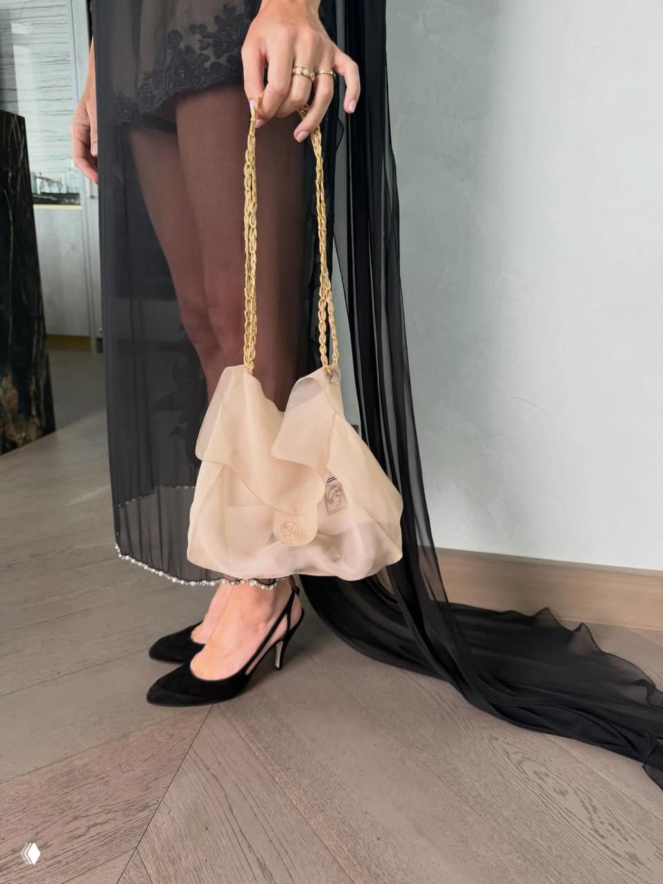 Рука держит небольшую светлую мягкую сумочку на тонкой цепочке; кадр акцентирует детали аксессуара и сочетание с прозрачной юбкой и туфлями.