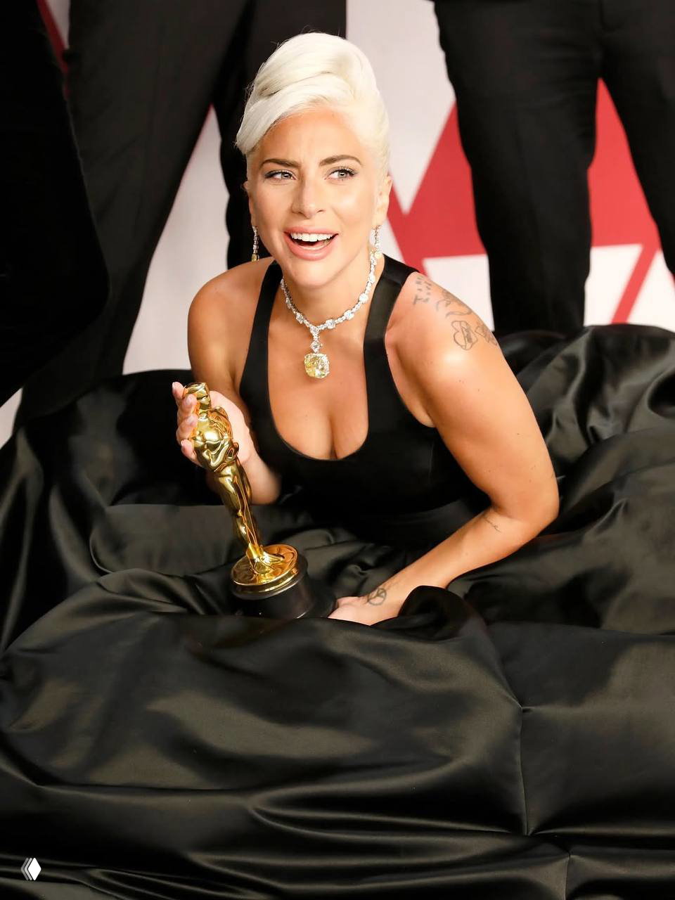 Цветная фотография на красной дорожке: женщина в чёрном вечернем платье держит статуэтку, яркая улыбка и крупные драгоценности.