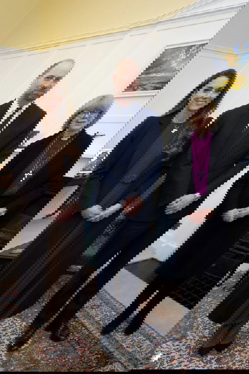 Групповое фото в помещении: женщина в коричневом образе, мужчина в тёмном костюме и представитель духовенства позируют в комнате с камином и ковром.