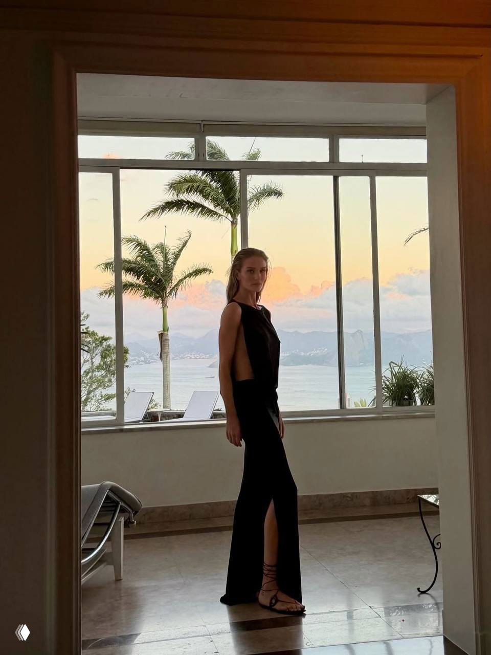 Женщина в длинном чёрном платье стоит у панорамного окна с видом на пальмы, море и мягкие закатные облака.