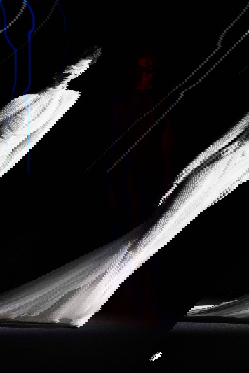 Белое облегающее кутюрное платье со скульптурными швами и длинным полупрозрачным шлейфом, модель в минималистичном образе на затемнённом фоне.