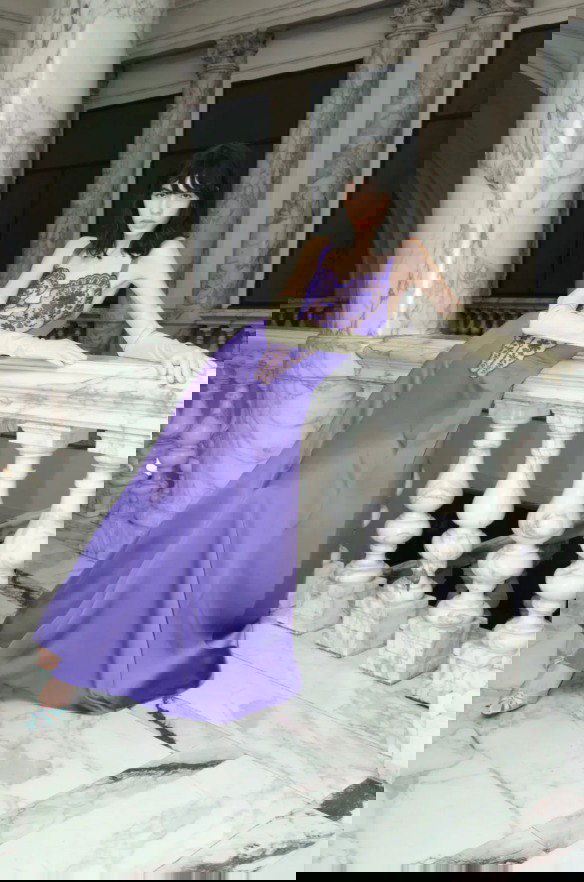 Модель в сиреневом атласном платье с кружевной вставкой опирается на мраморный парапет — элегантный вечерний образ из Pre‑Fall 2026.