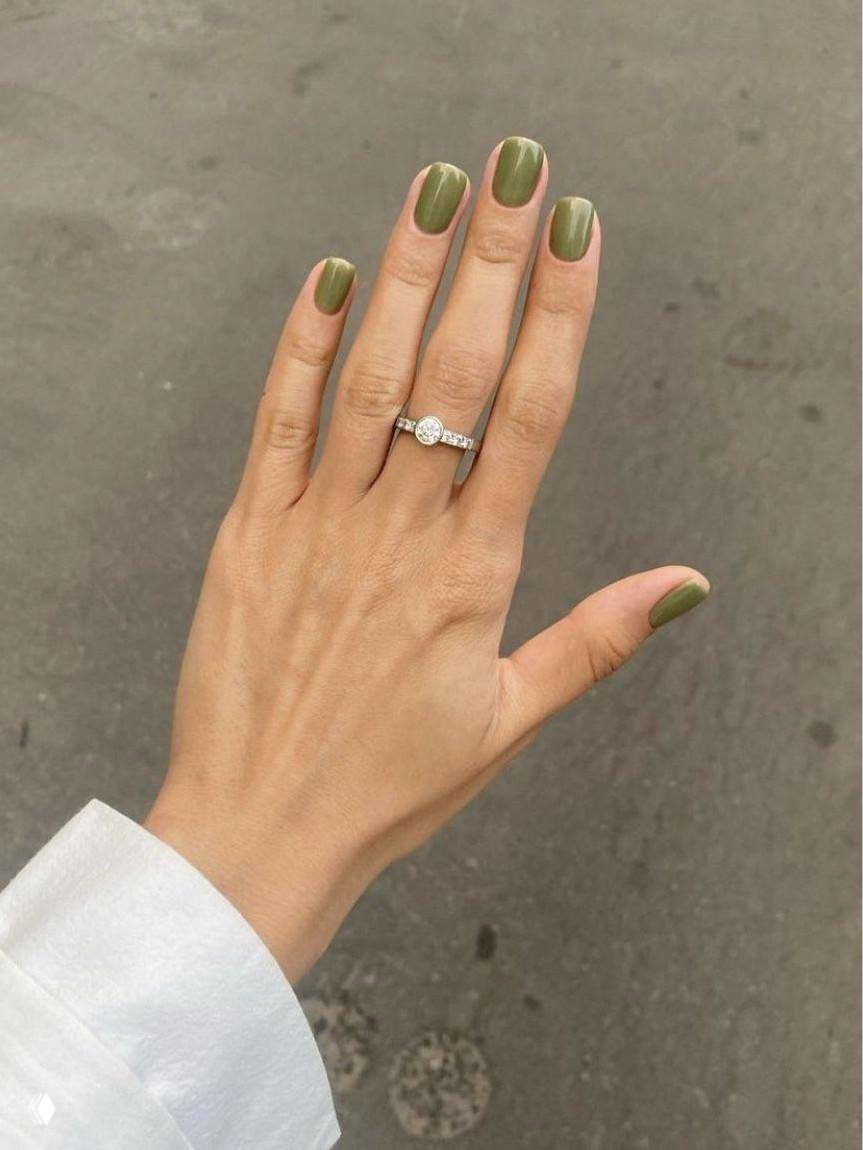 Рука с оливково-зеленым маникюром на фоне асфальта, аккуратная форма ногтей, натуральный оттенок кожи и простое украшение в виде кольца.