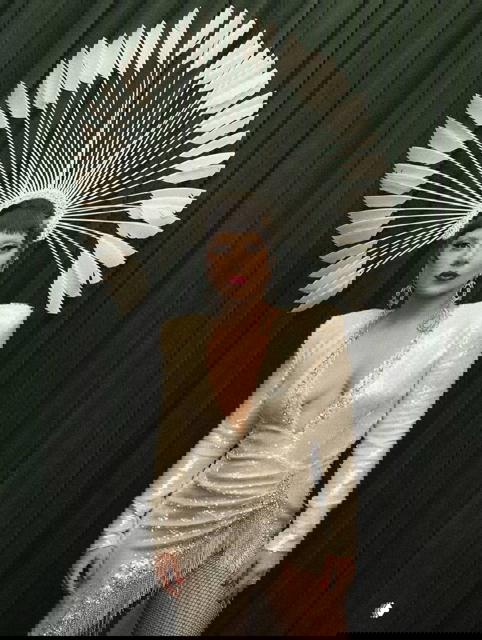 Женщина в светлом блестящем платье с глубоким декольте и высоким головным убором в виде веера, позирует на фоне тёмно‑зелёной ткани.