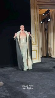 Модель идёт по подиуму в длинном светлом платье с драпировкой и в чёрном плаще на показе Giorgio Armani Haute Couture, атмосфера подиума и драматичное освещение.