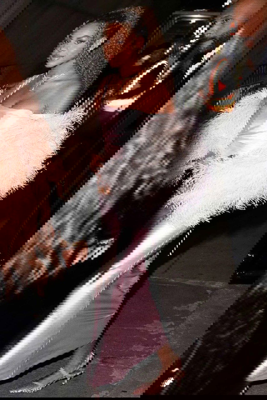 Полноформатный кадр: подросток идёт в длинном атласном платье и пушистом жакете, вечернее освещение и люди позади неё.