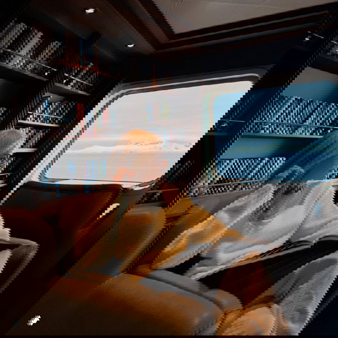 Интерьер каюты: человек в светлом свитере сидит в кресле у окна и смотрит на ледяной горизонт, на заднем плане полки с книгами и мягкий свет.