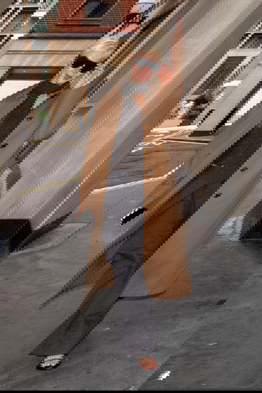 Женская фигура в коричневом пальто и солнцезащитных очках, длинное пальто и темные брюки, прохожие и архитектура на заднем плане.