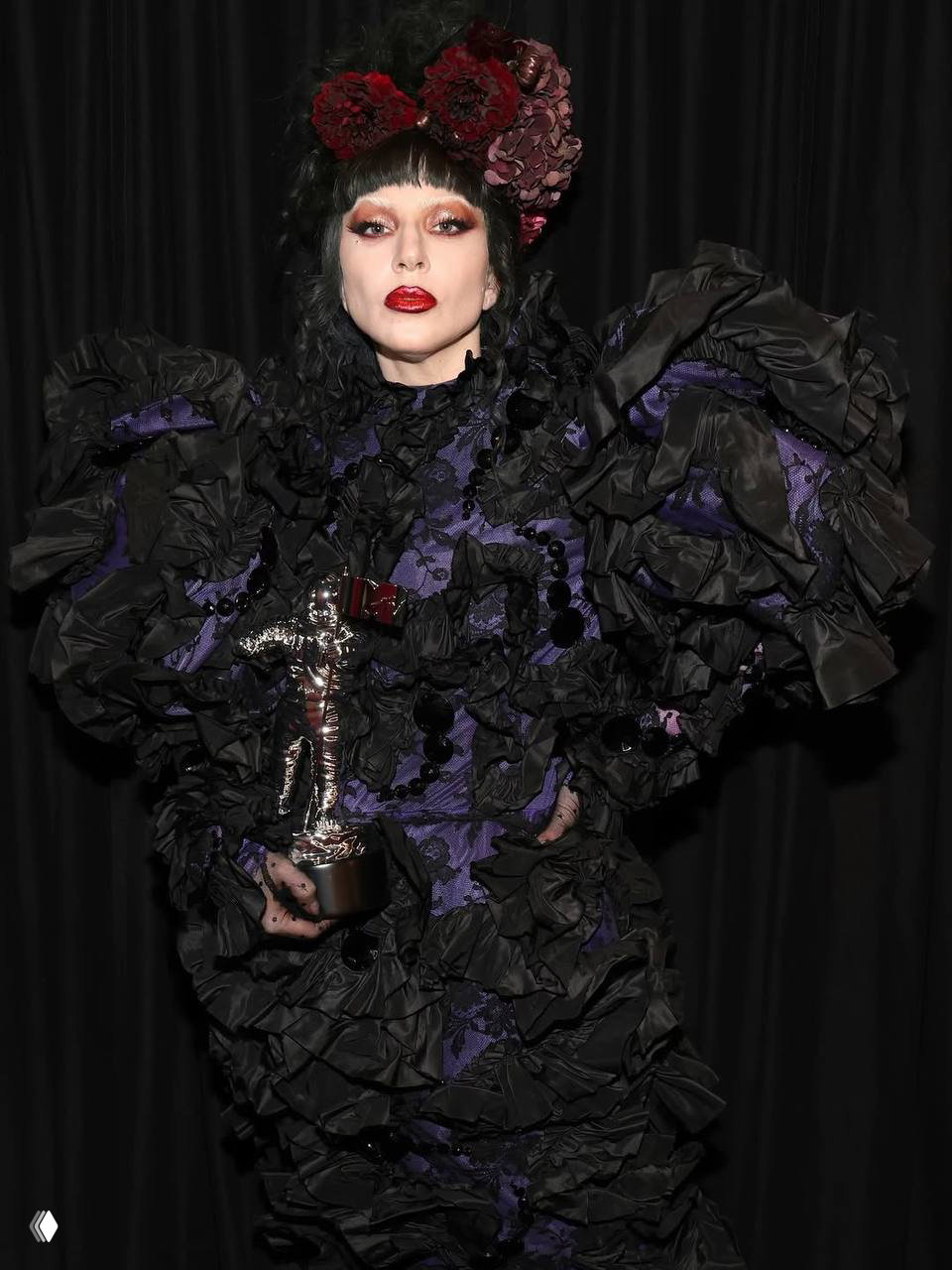 Портрет в театральном образе: женщина в сложном чёрном наряде с пышными рукавами и цветочными украшениями в причёске, держит награду в руках.