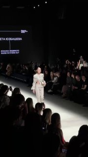 Модель в светлом костюме идёт по дорожке: аудитория занята съёмкой и наблюдением, атмосфера показа начинающих дизайнеров на Московской неделе моды.