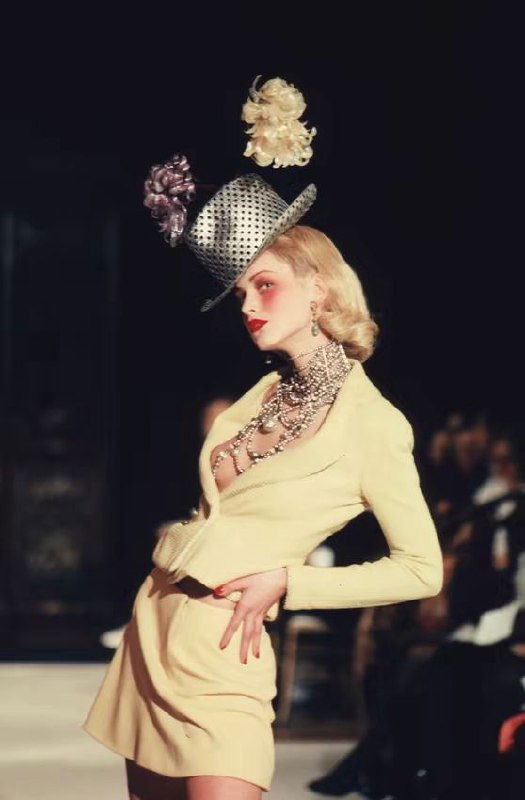 Модель в образе осенней коллекции Dior 1998: светло‑жёлтый жакет и мини‑юбка, высокая шляпа с цветком и многослойные жемчужные ожерелья на подиуме.