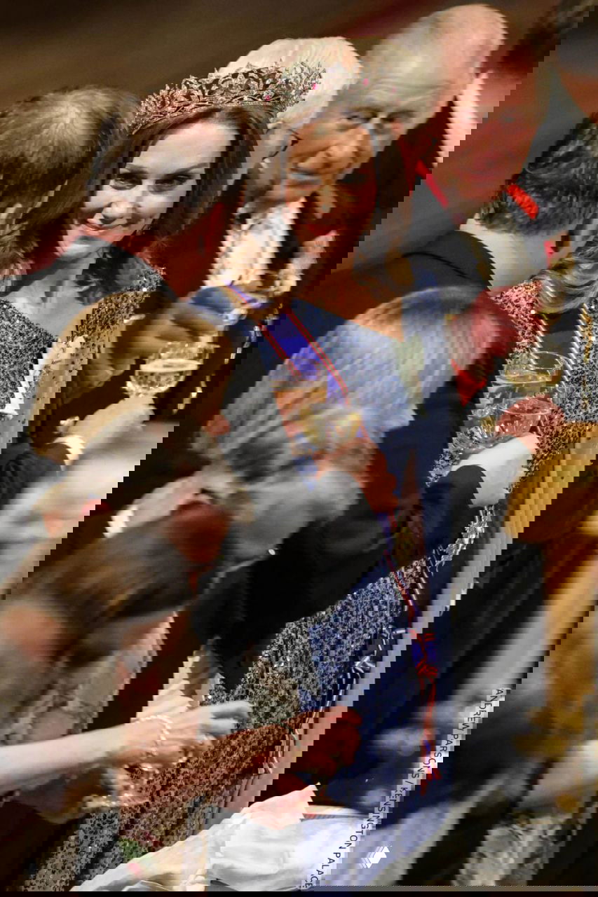 Крупный план женщины в блестящем платье и тиаре, она держит бокал и общается с гостями за праздничным столом в замке.