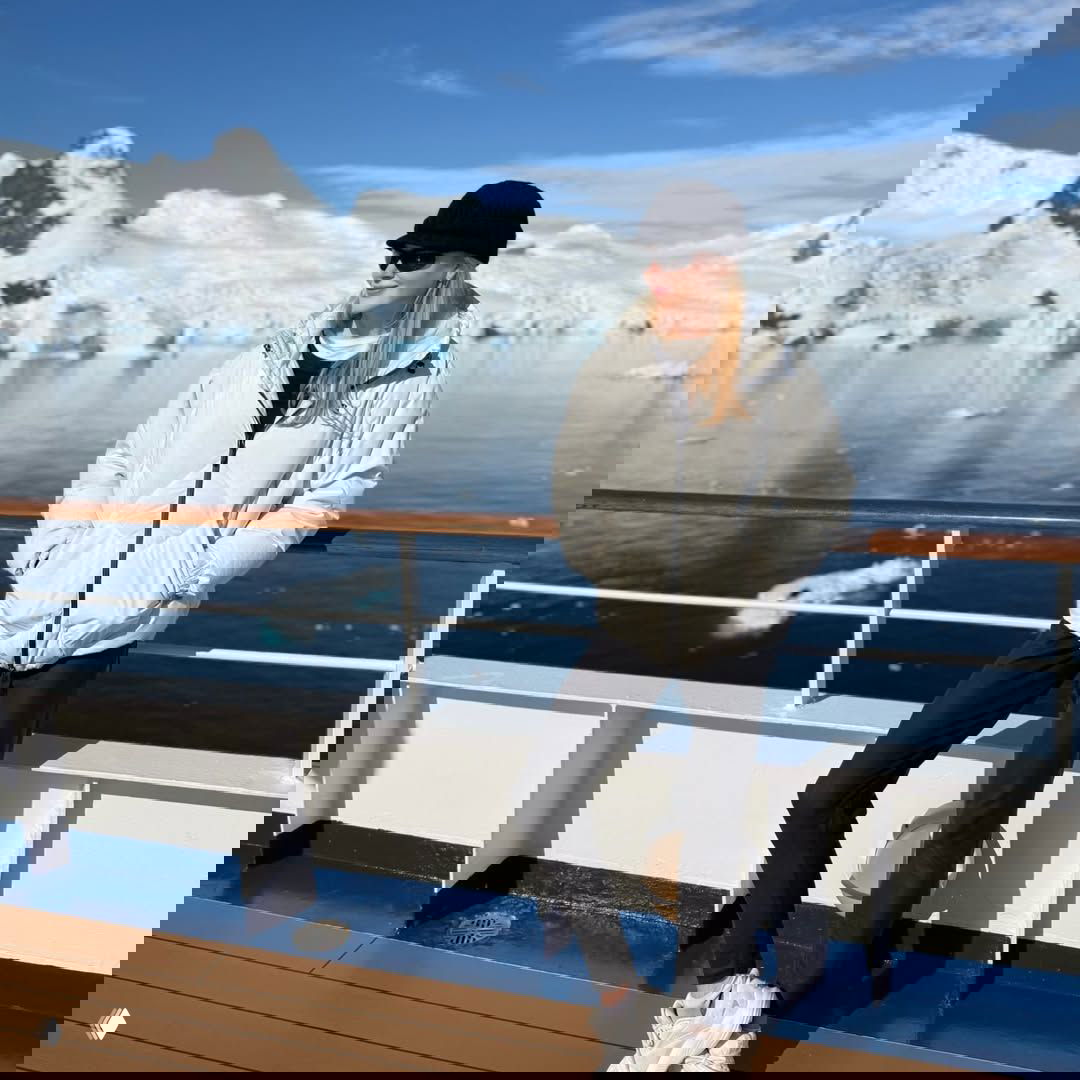 Пассажир на палубе корабля в светлой куртке позирует на фоне чистой воды и ледниковых фьордов — ясный день и зеркальная поверхность моря.