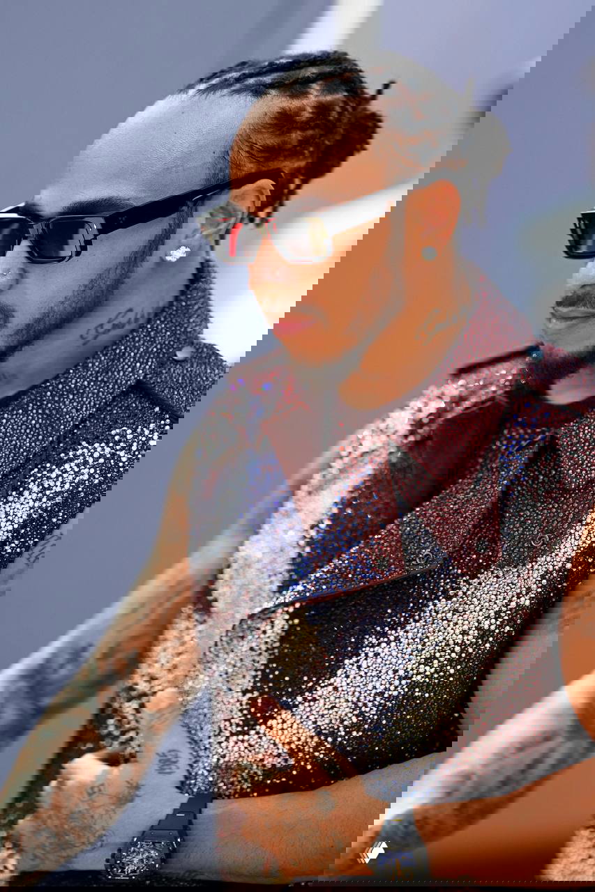 Крупный план Льюиса Хэмилтона в солнечных очках и сверкающем жилете; детали украшений и татуировок в кадре подчеркивают индивидуальность.