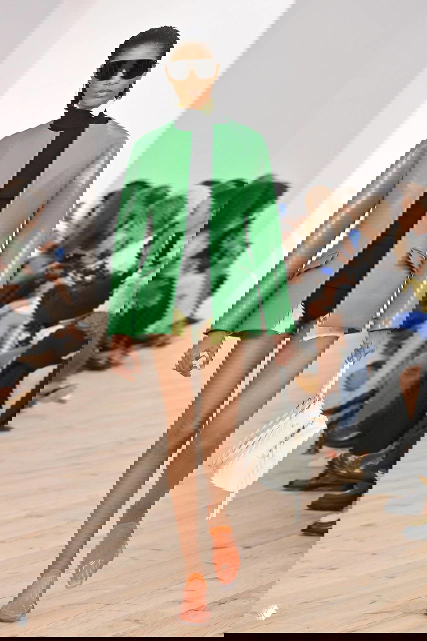 Показ: модель в коротком черном боди и ярко-зеленом жакете, прозрачная сумка в руке, пол и трибуны с публикой на фоне.