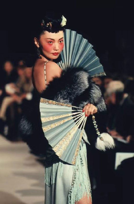 Модель с веером в руках, меховым манто и платьем в пастельных тонах на подиуме Dior 1998 — акцент на аксессуарах, позах и сценографии показа мод.