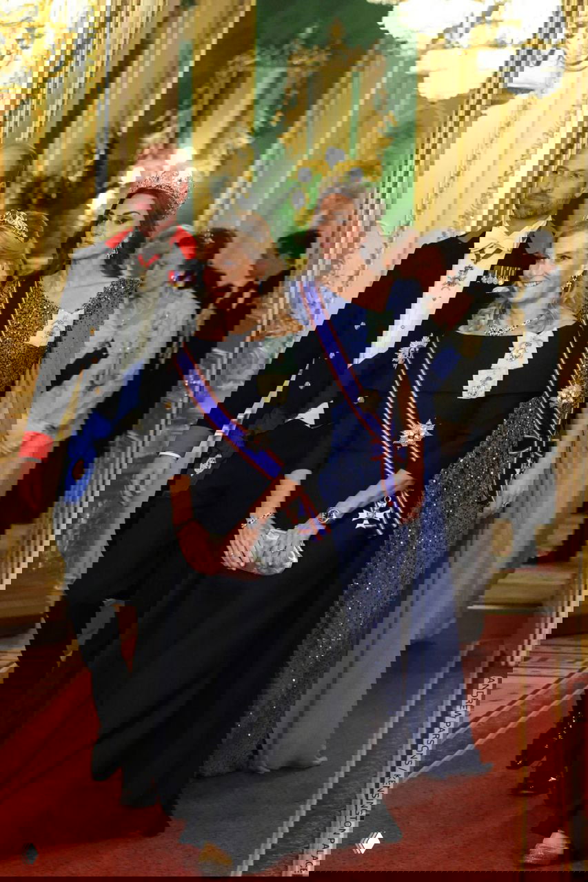 Несколько гостей в парадных костюмах и дамы в вечерних платьях идут по коридору с ковровой дорожкой и хрустальными люстрами.