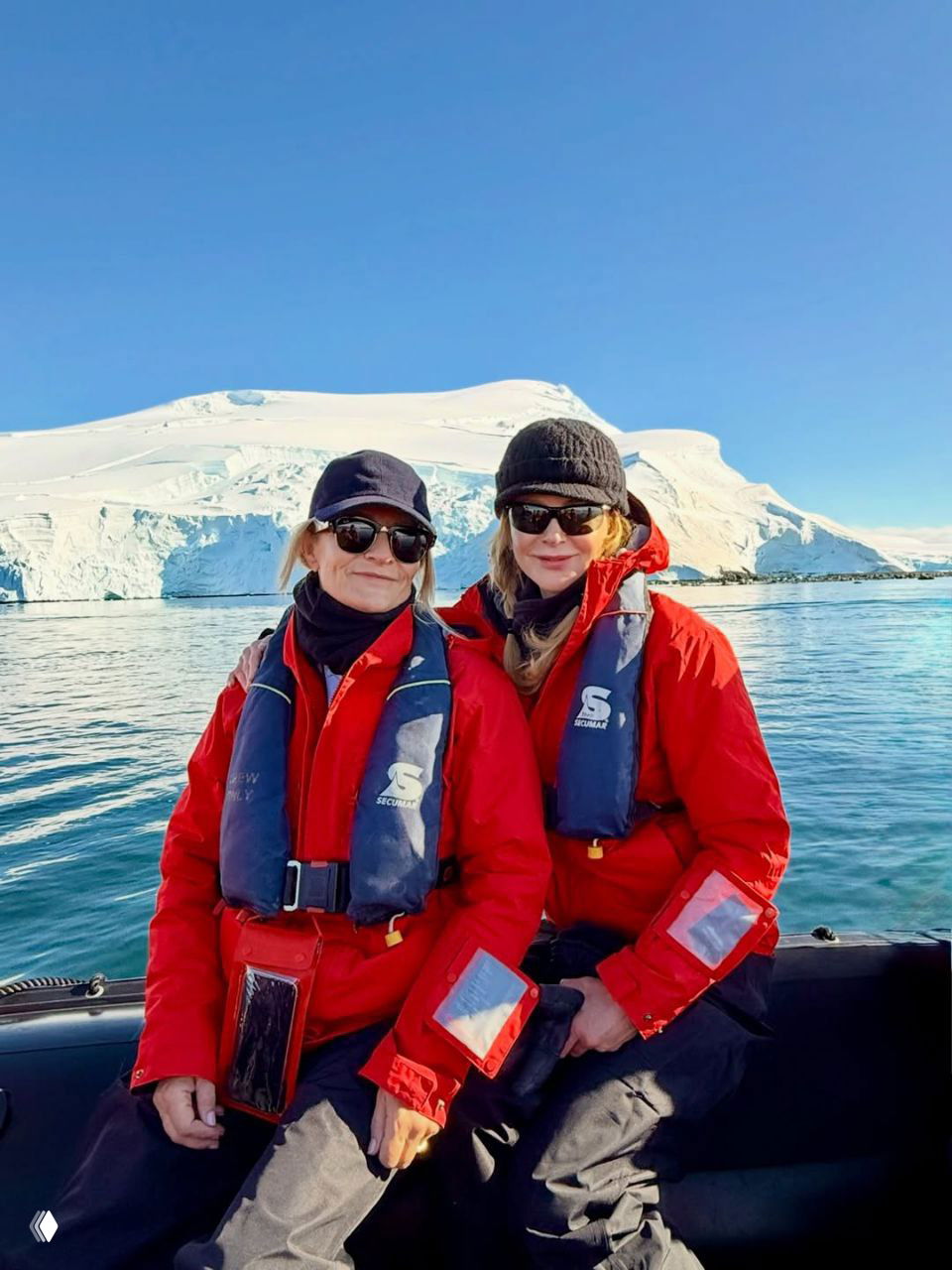 Два человека в спасжилетах и красных куртках сидят в небольшой лодке-зодиаке на спокойной воде у ледового массива в солнечный день.