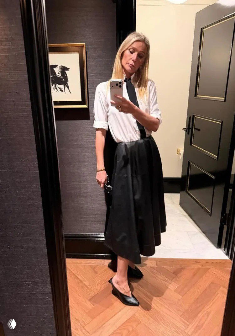 Гвинет Пэлтроу стоит в зеркале в белой рубашке и тёмной юбке с галстукоподобным акцентом, образ в стиле минималистичной элегантности на показе