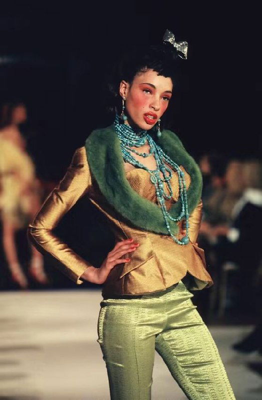 Модель в золотистом жакете и салатовых брюках на подиуме Dior 1998, многослойные бирюзовые бусы и выразительный макияж в стилизованной «азиатской» эстетике шоу.
