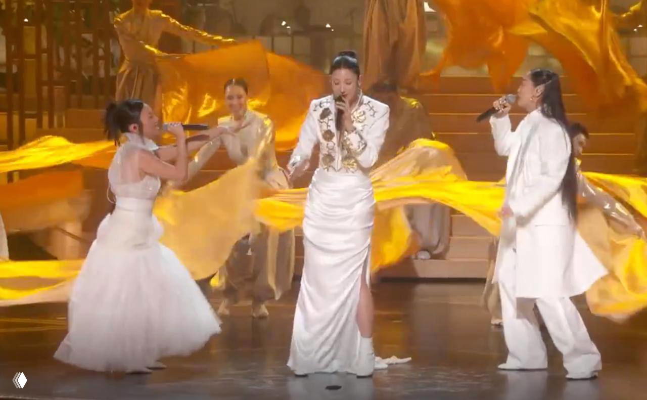 Музыкальное выступление на церемонии: несколько певиц в белых нарядах поют на фоне золотых развевающихся тканей и декораций.