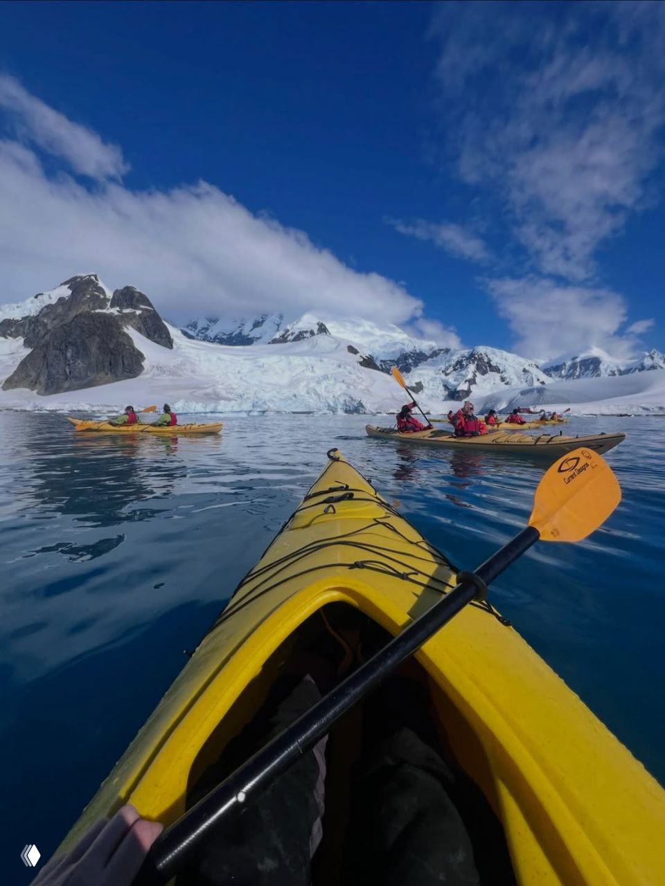 Вид от первого лица из жёлтого каяка: весло в кадре и группа каяков на зеркальной воде у заснеженных гор и айсбергов в ясную погоду.