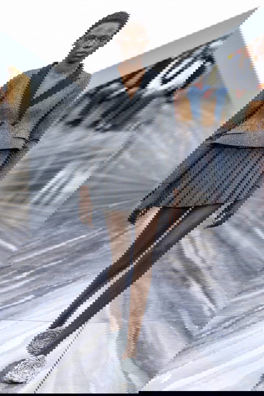 Модель в графичном сером жакете и плиссированной мини-юбке идет по подиуму; внизу текстурные туфли-аксессуары похожие на цветы.