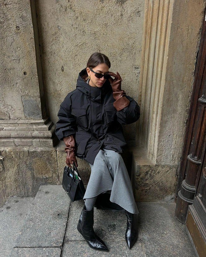 Стритстайл: девушка в чёрном объёмном пуховике и длинных коричневых кожаных перчатках, сидит у каменной стены с сумкой — городской зимний образ.