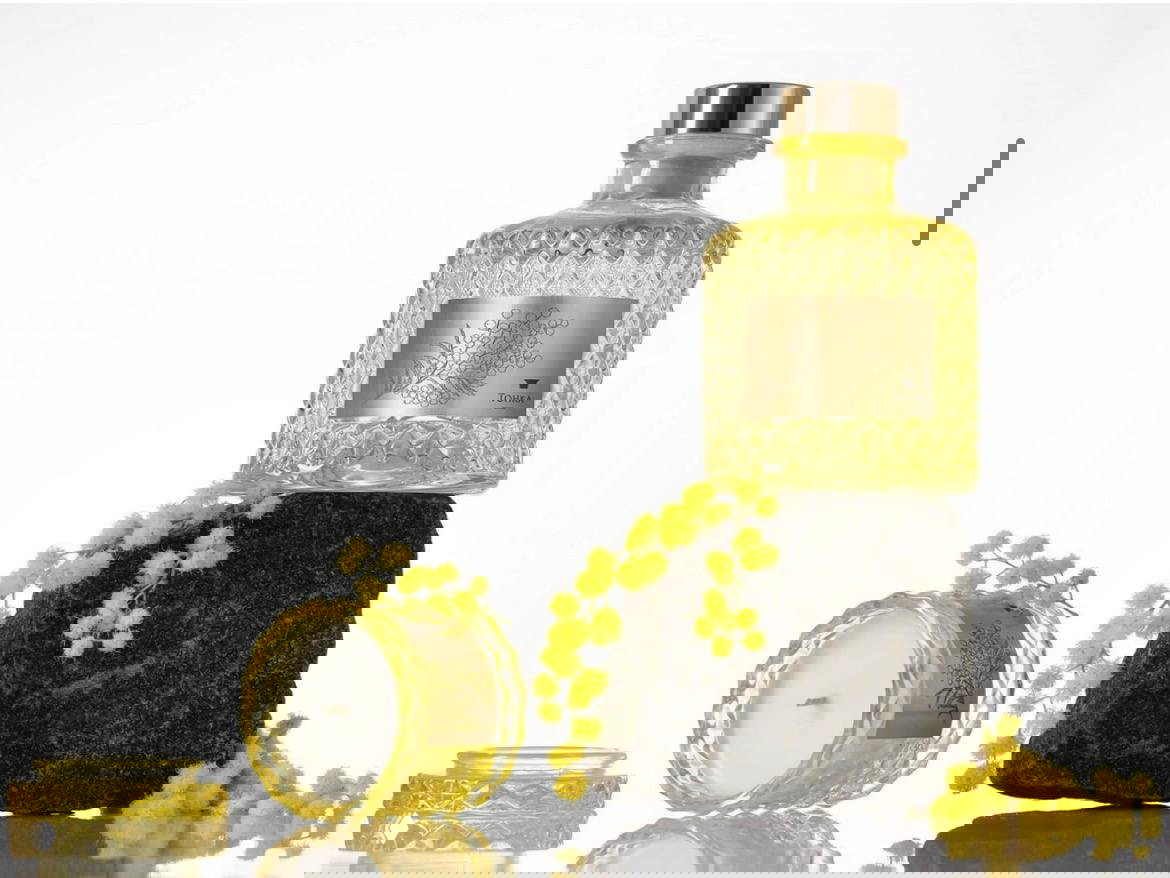 Жёлтая хрустальная свеча и флакон аромата Tonka Parfum на камне в стилизованной студийной композиции с веточками мимозы и отражениями.