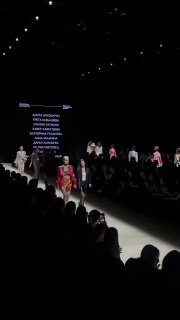 Финальная сцена показа: несколько моделей выстроены на подиуме, крупный экран с перечнем имён дизайнеров, публика аплодирует в полумраке.
