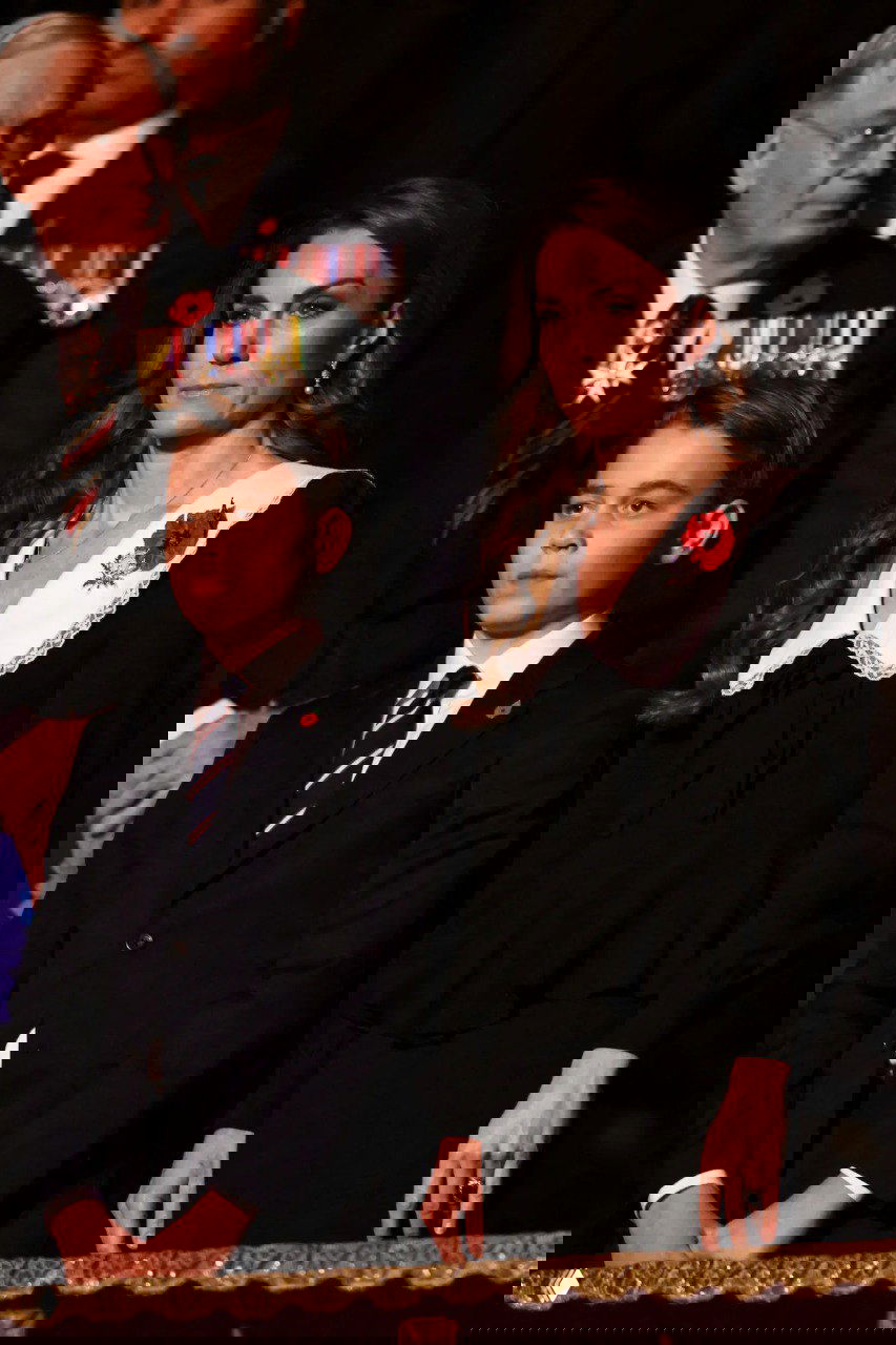 Они стоят в зале во время памятного концерта: строгие тёмные наряды, красный мак на лацкане и медали на фоне.