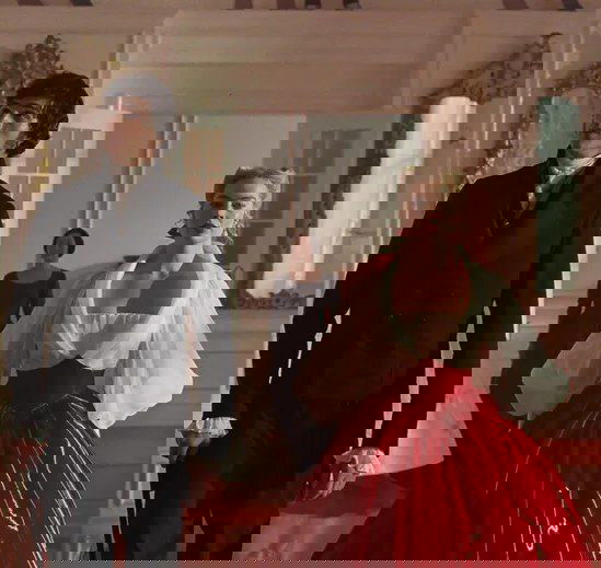 Пара персонажей в коридоре: женщина в ярком красном бальном платье и мужчина в темном костюме — кадровая сцена массового эпизода фильма.